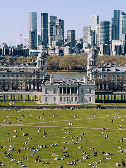 A panoramic view of Greenwich's skyline featuring modern glass skyscrapers and high-rise buildings, with construction cranes visible among the structures. In the foreground, a lush green park with wel