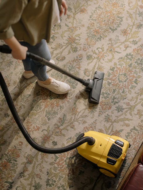 A person is vacuuming a traditional patterned area rug with a yellow vacuum cleaner, in a domestic setting. The individual is wearing beige shoes, blue jeans, and a beige jacket, and is holding the va
