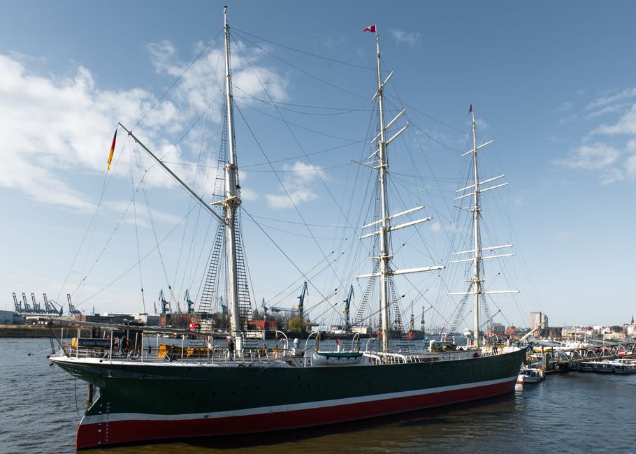 A large sailing ship docked at a harbor with three tall masts, rigging, and sails furled. The vessel has a dark green hull with a red and white stripe along the waterline, visible windows along the side, and a wooden deck. The background shows a clear blue sky with some clouds, industrial port structures and cranes in the distance, and a calm water surface reflecting the ship. This scene depicts a maritime setting, emphasizing the ship's clean, well-maintained appearance in the context of surface cleaning expertise, relevant to professional cleaning services provided by Carpet Cleaners Greenwich.