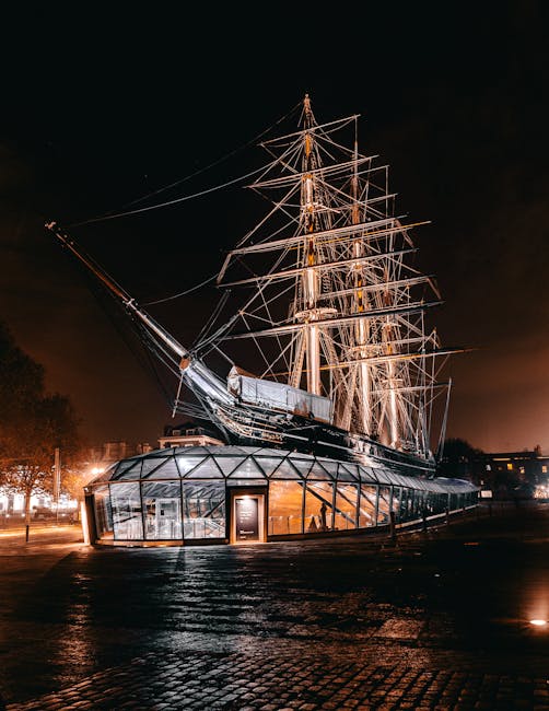 The image features the historic Cutty Sark clipper ship illuminated at night in Greenwich, with its tall masts and intricate rigging prominently visible against a dark sky. The ship's hull is enclosed with a modern glass structure, reflecting the surrounding lights, and the wet cobblestone ground in the foreground adds to the atmospheric setting. This scene showcases the vessel's detailed wooden decks, metallic fittings, and the contrast between the illuminated ship and the night sky. Carpet Cleaners Greenwich specializes in surface cleaning and deep cleaning services for residential and commercial spaces, ensuring hygienic and well-maintained environments. The ship's well-lit appearance emphasizes the importance of thorough sanitisation and maintenance, aligning with the principles of domestic and commercial cleaning for optimal hygiene and surface care.