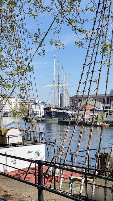 A large sailing ship docked at a harbor with three tall masts, rigging, and sails furled. The vessel has a dark green hull with a red and white stripe along the waterline, visible windows along the side, and a wooden deck. The background shows a clear blue sky with some clouds, industrial port structures and cranes in the distance, and a calm water surface reflecting the ship. This scene depicts a maritime setting, emphasizing the ship's clean, well-maintained appearance in the context of surface cleaning expertise, relevant to professional cleaning services provided by Carpet Cleaners Greenwich.