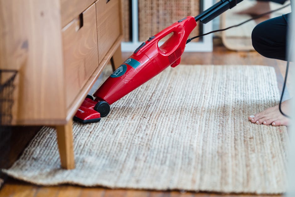 A professional cleaning technician from Carpet Cleaners Greenwich is performing deep surface cleaning in a living room. The room features a beige fabric sofa with matching cushions, a light-colored wo