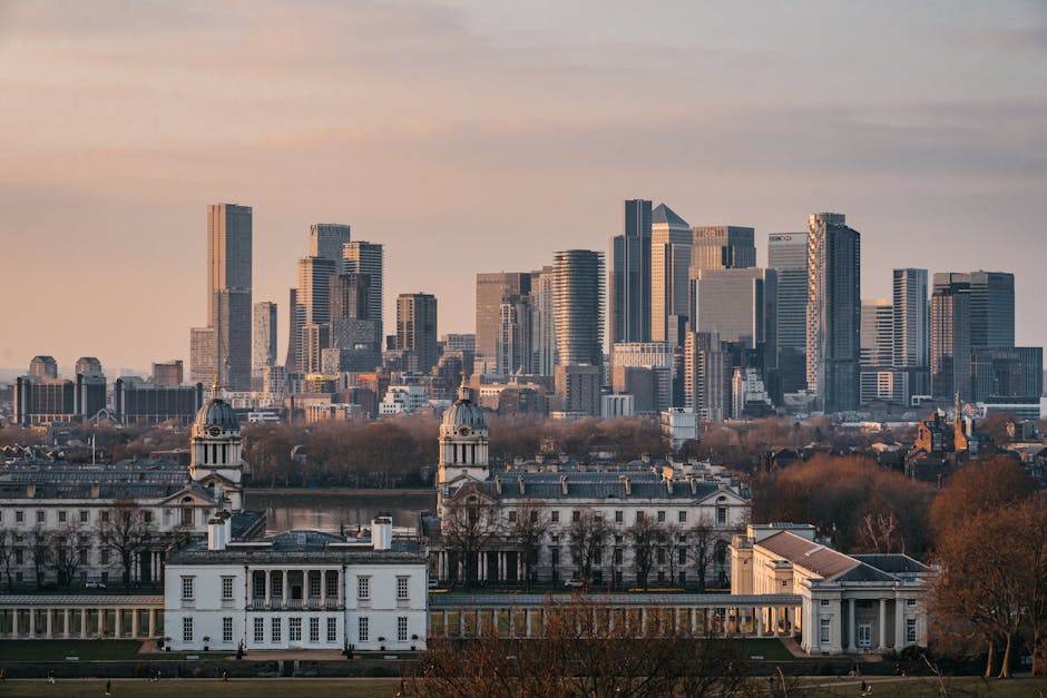 A cityscape of Greenwich SE10 featuring a cluster of modern skyscrapers with glass facades reflecting the sky, contrasted by historic white buildings with classical architecture, domed towers, and copper roofs in the foreground. The scene is captured during late afternoon or early evening with soft natural lighting, and the area is surrounded by leafless trees indicating autumn or winter. The image embodies a blend of urban development and preserved heritage, representative of Greenwich's skyline, aligned with the services provided by Carpet Cleaners Greenwich in the context of surface cleaning and deep cleaning discussions.