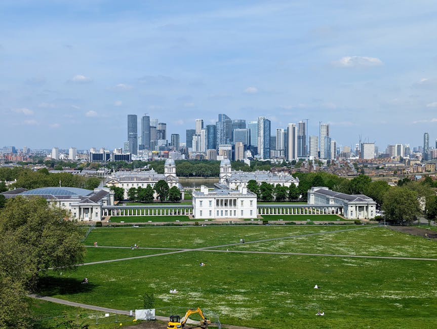 A wide view of Greenwich Park showing the well-maintained green lawn and pathways, with the historic Queen's House white building featuring classical architecture, columns, and a domed roof in the foreground. In the background, the modern skyline of Greenwich with tall glass skyscrapers rises against a partly cloudy sky, highlighting the contrast between historical and contemporary city elements. The area appears clean and orderly, with some small groups of people visible enjoying the outdoor space, and construction equipment seen near the edge of the grass. Bright daylight illuminates the scene, emphasizing the vivid green of the park and the reflective surfaces of the surrounding buildings. The image is associated with professional cleaning and maintenance services provided by Carpet Cleaners Greenwich for residential and commercial spaces, emphasizing surface cleaning, sanitisation, and deep cleaning solutions in the Greenwich SE10 area.