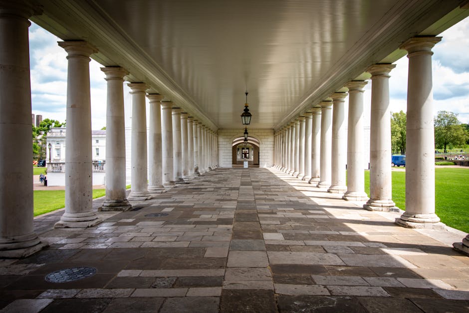 A covered outdoor walkway supported by a series of tall, white, classical-style columns with rounded bases, extending into the distance under a beige ceiling. The floor consists of large stone tiles arranged in a pattern, appearing clean and well-maintained. Classic lantern-style hanging lights are suspended from the ceiling at regular intervals. The area is bathed in natural daylight, with lush green grass and trees visible on either side of the walkway, and a partly cloudy sky in the background. This setting exemplifies surface cleaning and maintenance of historic architectural features, aligning with the theme of domestic cleaning and surface sanitisation, as provided by Carpet Cleaners Greenwich.