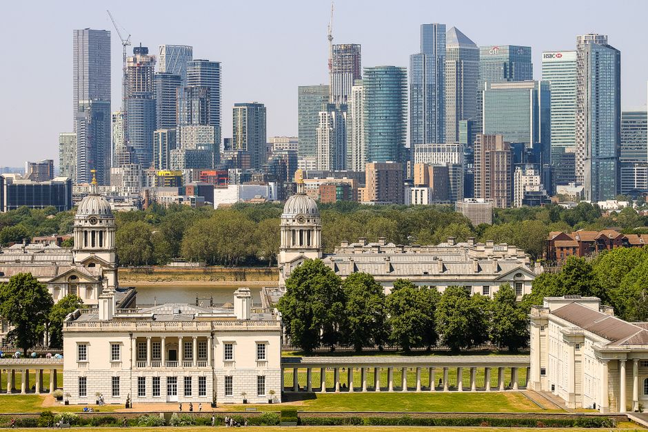 A panoramic view of a modern city skyline featuring tall skyscrapers with glass facades and construction cranes, set against a backdrop of a partly cloudy sky. In the foreground, a lush green park wit