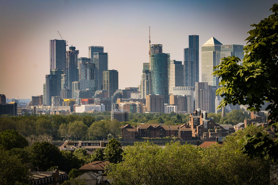 A panoramic view of the Greenwich city skyline under a partly cloudy sky, featuring tall modern office and residential buildings with glass facades reflecting ambient light. In the foreground, there i