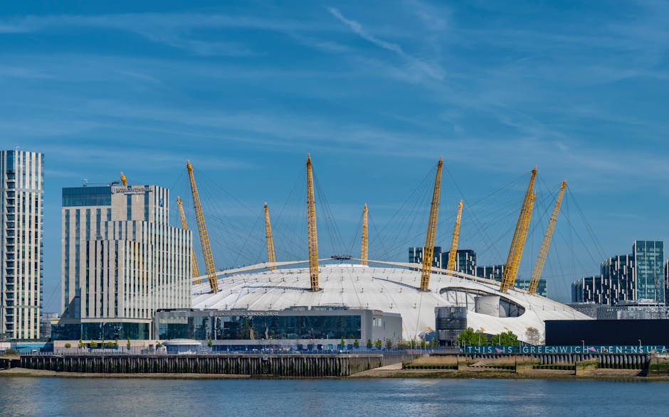 A panoramic view of the O2 Arena in Greenwich, London, featuring its large white dome roof supported by yellow tensile pillars and surrounded by modern high-rise office buildings. The waterfront area is visible in the foreground with calm water reflecting the structures and a clear sky overhead, highlighting the urban landscape and iconic architecture of the Greenwich area. This image captures the clean and well-maintained environment typical of the vibrant district, with no visible dirt or clutter.
