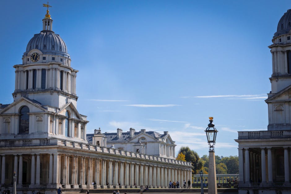 A panoramic view of historic white stone buildings with domed roofs and classical architectural details in Greenwich, under a clear blue sky. In the foreground, a black ornate street lamp stands next to the buildings. The area appears well-maintained and clean, with a few visitors walking along the paved paths, and lush green trees are visible in the background. This scene exemplifies the tidy and hygienic environment associated with professional domestic cleaning services such as those offered by Carpet Cleaners Greenwich, emphasizing surface cleaning and maintenance in a historic setting.