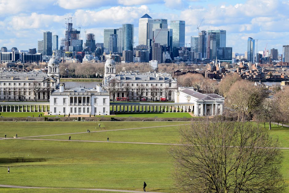 A panoramic view of the O2 Arena in Greenwich, London, featuring its large white dome roof supported by yellow tensile pillars and surrounded by modern high-rise office buildings. The waterfront area is visible in the foreground with calm water reflecting the structures and a clear sky overhead, highlighting the urban landscape and iconic architecture of the Greenwich area. This image captures the clean and well-maintained environment typical of the vibrant district, with no visible dirt or clutter.